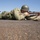 A person in WWII-era U.S. Army uniform is lying prone on a shooting range surface, aiming a rifle downrange. The individual is wearing period-accurate olive drab clothing, an M1 helmet, and web gear with ammunition pouches. The setting appears to be a desert shooting range, consistent with a historical reenactment or marksmanship demonstration.