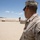 A U.S. Marine in desert MARPAT camouflage uniform is firing a pistol at an outdoor shooting range in an arid desert environment. The shooter is in a two-handed grip stance, aiming downrange at a row of targets mounted on a berm. The setting appears to be a military training facility, likely in a southwestern U.S. desert location.
