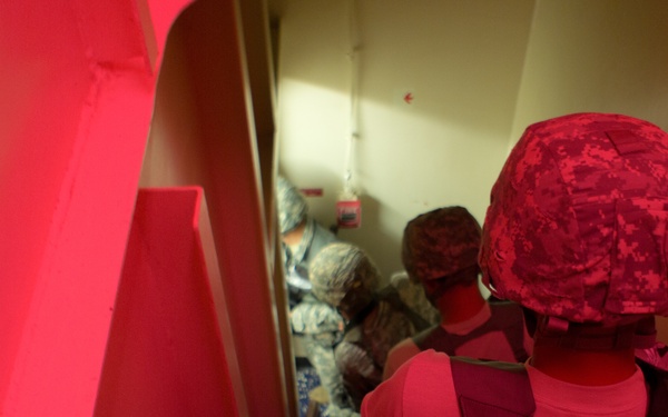 The image shows multiple individuals in military or tactical gear moving through a narrow corridor or stairwell. The scene is dominated by a strong red light source on the left side, casting a red tint over much of the frame. The personnel appear to be conducting a tactical movement or building clearance exercise.