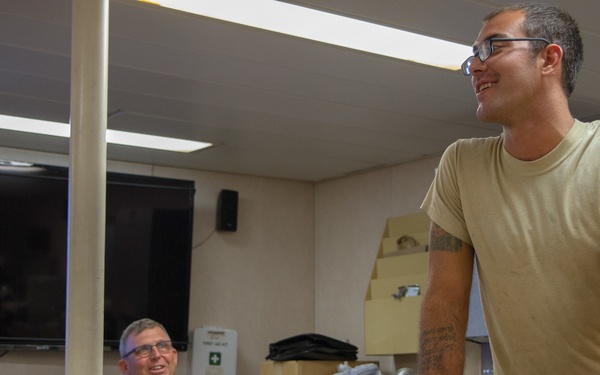 Two military personnel are in an indoor facility, likely a briefing or work room. One soldier in full U.S. Army camouflage uniform is seated at a table, while another in a tan t-shirt and glasses stands at the table handling what appears to be a weapon system or military equipment. Both individuals appear relaxed and are smiling.