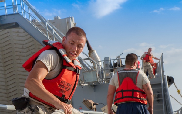 Military personnel in U.S. Army camouflage uniforms and red Stearns work vests are performing line-handling duties on the deck of a military vessel. The foreground soldier is actively working with a thick mooring rope around a deck bollard. Additional personnel are visible on stairs and the upper deck in the background.