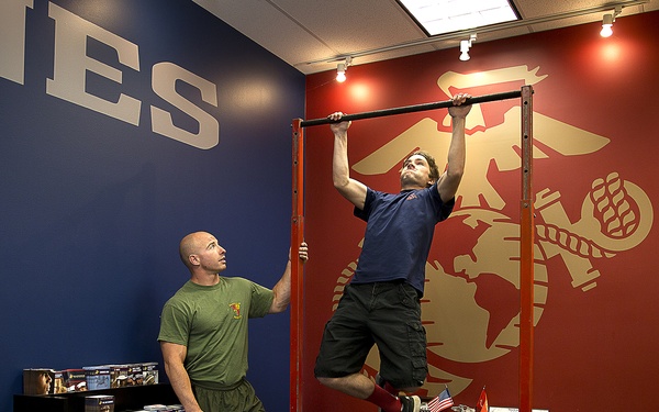 A man in a blue shirt and black shorts performs pull-ups on a red metal pull-up bar station inside what appears to be a U.S. Marine Corps recruiting office. A second man in an olive green military-style t-shirt and shorts observes and spots him. The room features the USMC Eagle, Globe, and Anchor emblem on a red wall, along with the word 'MARINES' on a blue wall.