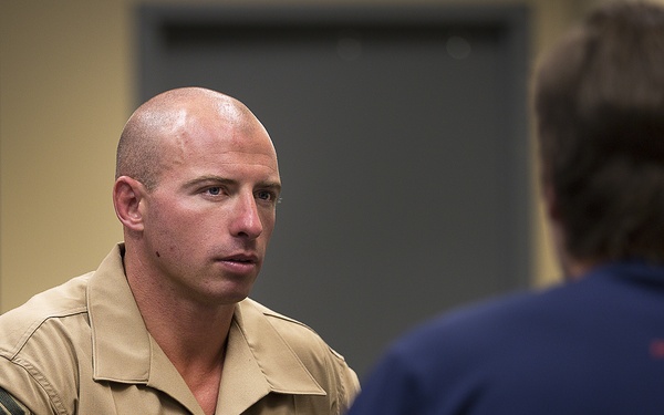 A U.S. Marine Corps sergeant named Leifheit, wearing the tan Service 'C' uniform with rank insignia and service ribbons, is seated and engaged in a face-to-face conversation with another individual whose back is to the camera. The setting appears to be an indoor office or meeting room with red walls and a gray door visible in the background. The interaction has the appearance of a formal or semi-formal interview or counseling session.