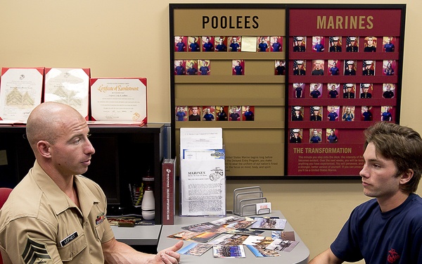 A U.S. Marine Corps recruiter in tan service uniform is meeting with a young civilian male at a small round table in a recruiting office. The office displays a 'Poolees and Marines' tracking board showing photos of recruits in various stages of enlistment, along with certificates and Marine Corps materials. This appears to be a standard recruiting consultation or enlistment interview.