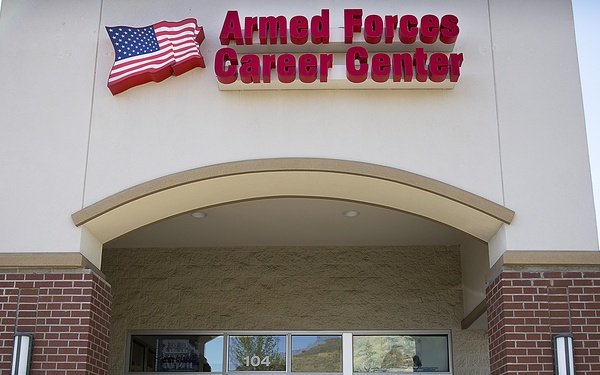 Two individuals are walking outside an Armed Forces Career Center, a military recruitment facility located in suite 104. One person is wearing a U.S. Marine Corps dress uniform, while the other is dressed in civilian clothing. The building features brick accents, an American flag logo, and window decals for the Marines and Air Force branches.