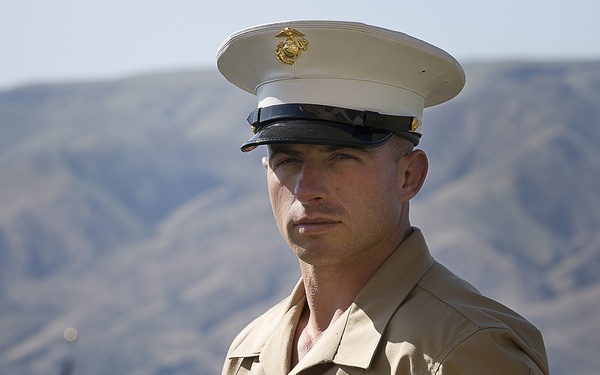 A U.S. Marine in Service Charlie uniform (tan short-sleeve shirt with white cover/cap) stands at attention or at ease outdoors. He displays rank insignia on his right sleeve consistent with a Staff Sergeant (E-6) and wears multiple service ribbons on his chest. The background features arid, hilly terrain and a partially visible American flag on a flagpole.