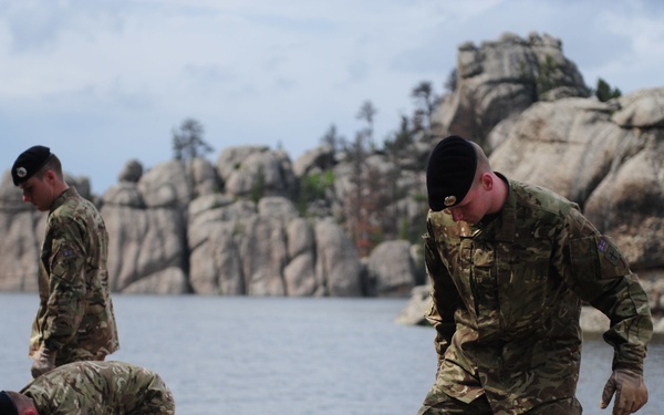 Several military personnel in camouflage uniforms and black berets are performing manual labor or maintenance work near a lakeside area. They appear to be handling wooden planks or boards on a sandy/gravelly shore. The backdrop features a scenic lake with distinctive granite rock formations, consistent with the Black Hills region of South Dakota, possibly Sylvan Lake.