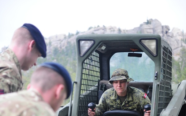 Military personnel from at least two different nations are engaged in a construction or engineering task involving a skid-steer loader. One soldier wearing Canadian Armed Forces CADPAT camouflage uniform with a Canadian flag patch operates the machine, while others in British MTP (Multi-Terrain Pattern) uniforms with blue berets work on the ground. The setting appears to be a rocky, forested terrain, suggesting a joint military exercise or field engineering operation.