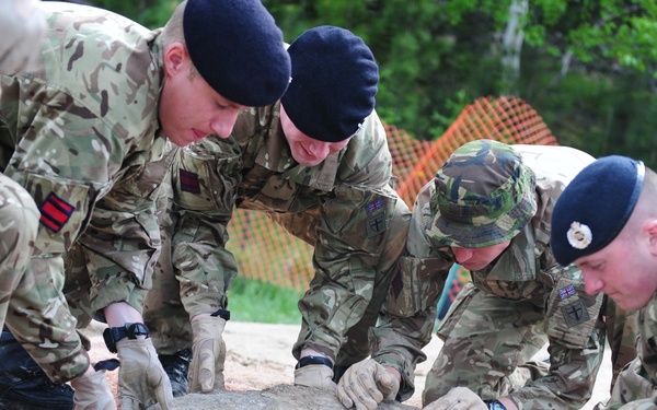 A group of military personnel in camouflage uniforms are collaboratively moving or positioning a large flat stone or boulder on a sandy/gravelly surface. The soldiers appear to be from British or NATO-affiliated forces, identifiable by their uniforms, berets, and shoulder patches. The activity appears to be a physical training exercise or construction/engineering task conducted outdoors.