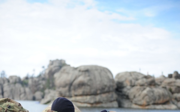 Three military personnel in British MultiTerrain Pattern (MTP) camouflage uniforms are working together to lift or move a large flat stone or concrete slab near a rocky waterfront. The soldiers appear to be engaged in a physical training exercise or engineering/construction task. The setting features distinctive rounded granite boulders and a body of water in the background.