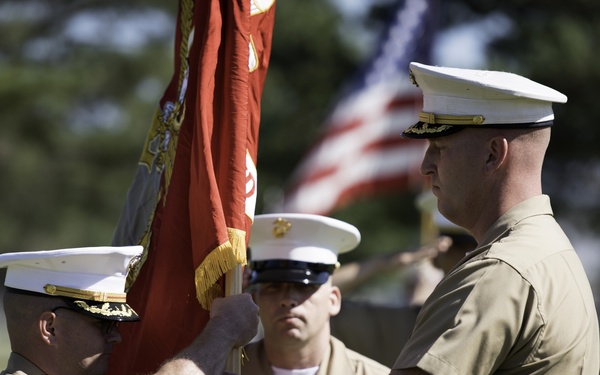 Two U.S. Marine Corps officers are participating in a formal change of command ceremony, passing a red unit guidon or colors between them. A third Marine stands at attention in the background, with an American flag and additional personnel visible in the distance. The ceremony is taking place outdoors on a grassy field in daylight.