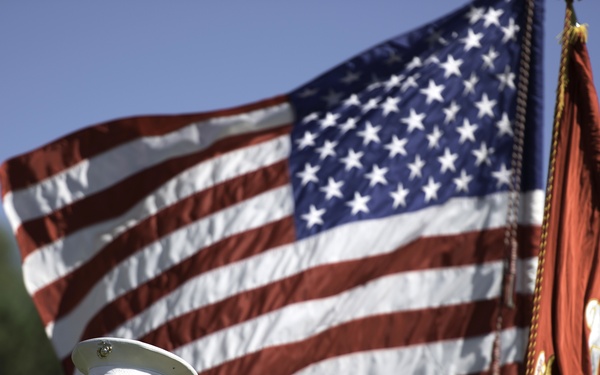A U.S. Marine Corps officer in service uniform renders a salute in front of a large American flag and a U.S. Marine Corps flag. The officer is wearing a white cover (cap) with gold insignia and a tan service uniform adorned with multiple service ribbons. The setting appears to be an outdoor military ceremony on a clear, sunny day.