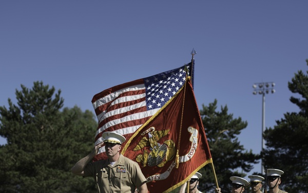 A United States Marine Corps color guard ceremony is taking place on a grass field under clear blue skies. A senior Marine officer in service uniform renders a salute while a color guard team carries the American flag and the USMC organizational flag. Additional Marines in formation are visible in the background, also at salute.