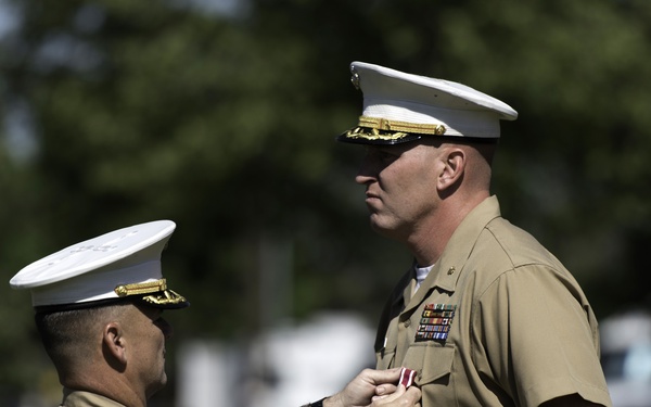 Two U.S. Marine Corps officers are participating in a military decoration ceremony outdoors. One officer is pinning a medal or award onto the chest of a taller officer who stands at attention. Both are wearing the standard Marine Corps Service 'Charlie' uniform with white covers (caps).
