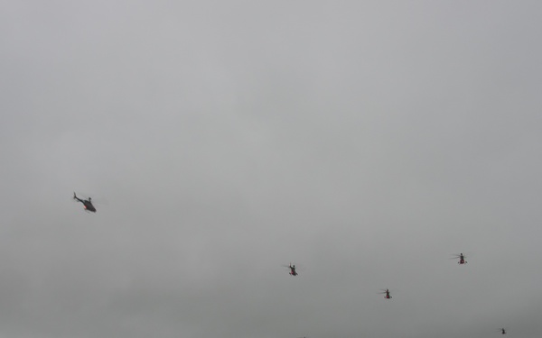 A military ceremony or commemoration event is taking place on a large open grass field, featuring historical reenactors in 18th-century colonial uniforms positioned beside a period cannon in the foreground. A large formation of modern military personnel in uniform stands in rows across the field, while multiple military helicopters fly in formation overhead against an overcast sky. The scene juxtaposes historical and contemporary military elements, suggesting an anniversary or commemorative military event.