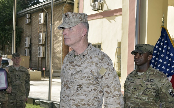 A military ceremony is taking place outdoors at what appears to be a forward operating base or military installation. A U.S. Marine officer in desert MARPAT camouflage is being presented with what appears to be a medal or award by a U.S. Army soldier. Several other military personnel are present, along with an American flag and a red general officer flag bearing two stars.