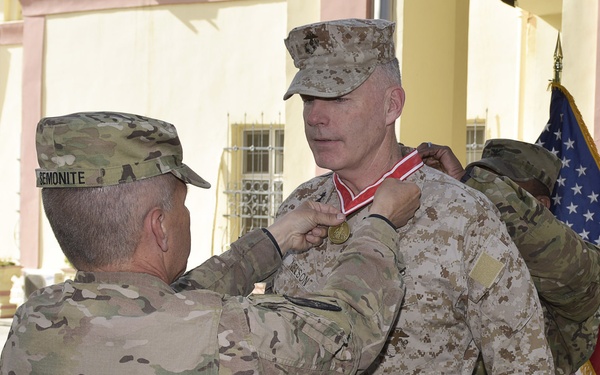 A military award ceremony is taking place outdoors, where a soldier in Army OCP (MultiCam) camouflage is pinning a medal with a red ribbon onto a Marine in MARPAT desert camouflage uniform. A third service member assists from behind. An American flag is visible in the background.