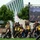 2015 Department of Defense Warrior Games Opening Ceremony A group of athletes wearing matching black and gold uniforms, some in wheelchairs, are gathered at what appears to be an opening ceremony for a military adaptive sports competition. They are carrying a white flag and moving as a team delegation, with a large LED display screen visible in the background. The setting appears to be an outdoor venue with a modern architectural building visible behind the trees.