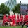 2015 Department of Defense Warrior Games Opening Ceremony A group of individuals wearing matching red and white 'Marines' athletic uniforms are participating in what appears to be a formal parade or ceremonial procession. The group includes both ambulatory participants and those using wheelchairs, along with at least one service dog wearing a yellow vest. A large LED display screen is visible in the background, along with a modern architectural building and trees.