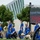 2015 Department of Defense Warrior Games Opening Ceremony A group of individuals wearing matching blue athletic uniforms are participating in what appears to be a ceremonial procession or parade, likely associated with a Paralympic or adaptive sports event such as the Warrior Games or Invictus Games. Several participants are in wheelchairs, and at least two service dogs are present alongside the athletes. A large LED display screen is visible in the background, along with a modern glass-and-steel building.