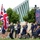 2015 Department of Defense Warrior Games Opening Ceremony A group of individuals wearing matching navy blue uniforms are participating in what appears to be a ceremonial parade or procession, led by a person carrying a large Union Jack (British flag). The group includes both ambulatory participants and individuals using wheelchairs, suggesting this is likely a military veterans or adaptive sports event such as the Invictus Games. A large LED screen in the background displays the procession, and official-looking observers are present.