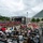 2015 Department of Defense Warrior Games Opening Ceremony A large outdoor ceremony is taking place in front of a distinctive modern building, identified by signage as the 'Warrior Games.' Hundreds of attendees are seated in folding chairs facing a large stage with a banner reading 'Welcome to The Games' and 'Warrior Games.' Many attendees are wearing black shirts reading 'Family Program - Team Army,' with other groups in red, blue, and black attire.