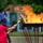 2015 Department of Defense Warrior Games Opening Ceremony A ceremonial torch lighting is taking place outdoors, with a civilian in a red polo shirt holding a torch to ignite a large cauldron-style flame bowl supported by red metal legs. A military service member in olive dress uniform and garrison cap stands alongside, appearing to assist or oversee the ceremony. Flags and trees are visible in the background, suggesting a formal commemorative or patriotic event.