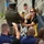 2015 Department of Defense Warrior Games Opening Ceremony A military ceremony is taking place outdoors where a uniformed Marine (facing away from camera) is handing or receiving a lit torch to/from a woman wearing a black and yellow athletic polo shirt and sunglasses. An audience of seated attendees is visible in the foreground, with several people photographing the event. Additional military personnel in various uniforms are visible in the background near a staircase.