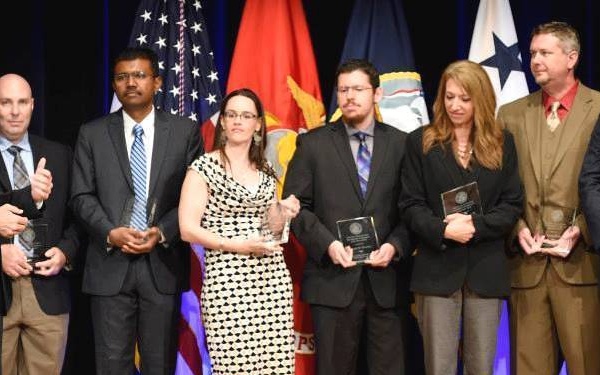 A group of approximately eleven individuals are standing on a stage during what appears to be a formal awards ceremony. Several recipients are holding plaques or awards, with military and government flags visible in the background. The setting suggests an official government or military recognition event.