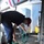 USS Blue Ridge operations A young person wearing a dark shirt, blue pants, and a red baseball cap is bent over coiling or uncoiling a green garden hose on a textured non-slip deck surface. The setting appears to be an industrial or maritime environment, indicated by the gray metal equipment, structural supports, and anti-slip flooring. A yellow container and gray electrical or mechanical cabinets are visible in the background.