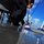 USS Blue Ridge operations The image shows personnel in dark uniforms scrubbing a wet deck surface using blue-handled push brooms, taken from a low ground-level angle. The setting appears to be the deck of a naval vessel, with a submarine or ship visible in the background. The dramatic low-angle perspective emphasizes the water pooling on the deck and the cleaning activity in progress.