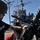 USS Blue Ridge operations Two U.S. Navy sailors are working with a mounted weapon system, likely an M2 .50 caliber machine gun, on the deck of a naval vessel. The sailor in the foreground is wearing a helmet, sunglasses, and a 'US NAVY' shirt with visible arm tattoos, while handling the weapon mount. A large naval warship with radar arrays and communication masts is visible in the background.
