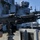 USS Blue Ridge operations A U.S. Navy sailor in combat gear is manning a mounted M240 machine gun on the deck of a naval vessel at sea. The sailor is wearing a Kevlar helmet and a black uniform with 'US' visible, and appears to be in a training or watch-standing posture. The ship's superstructure, radar equipment, and open ocean are visible in the background.