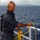 USS Blue Ridge operations A sailor in dark blue Navy working uniform stands on the deck of a naval vessel, handling a yellow rope or line near the ship's railing. Land masses and mountains are visible in the background across open blue ocean water. The individual appears to be performing routine deck seamanship duties.