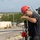 USS Blue Ridge operations Two individuals in dark military-style uniforms are positioned on an elevated platform or ship deck. The foreground figure wears a red hard hat and glasses and is handling equipment that includes a red spool of line/rope and a cylindrical tool. A coastal or port environment with buildings and vegetation is visible in the blurred background.