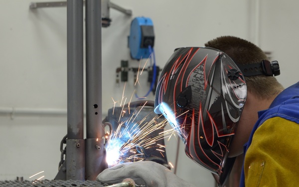 A welder is actively welding a metal structural component in what appears to be a workshop or training facility. The person is wearing full protective gear including a decorative welding helmet with red flame graphics, a blue work jacket with yellow leather sleeves, and white welding gloves. Bright sparks and arc light are visible at the welding point on a metal grating work surface.