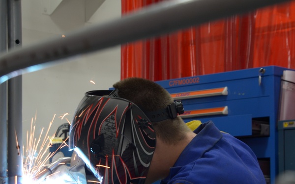 A welder is actively welding metal components on a perforated metal work table, with visible sparks and bright arc light. The person is wearing a decorative black and red welding helmet along with a blue work jacket and yellow welding gloves. The setting appears to be a professional or vocational workshop environment.
