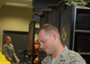A US Air Force Technical Sergeant, with last name partially visible as 'LDER', is examining yellow inflatable life vests or survival equipment in what appears to be a military equipment storage facility. The airman is wearing the standard Air Force digital camouflage uniform (ABU). A second uniformed service member is visible in the background.