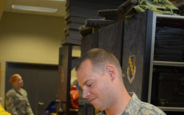 A US Air Force Technical Sergeant, with last name partially visible as 'LDER', is examining yellow inflatable life vests or survival equipment in what appears to be a military equipment storage facility. The airman is wearing the standard Air Force digital camouflage uniform (ABU). A second uniformed service member is visible in the background.
