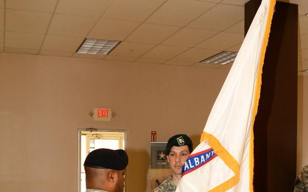 Two U.S. Army soldiers in ACU (Army Combat Uniform) are participating in what appears to be a formal military ceremony indoors. One soldier wearing a green beret (name tape reads 'COLEMAN') is holding a white guidon/flag with gold fringe bearing the word 'ALBANY', while a second soldier in a black beret faces him. The setting appears to be a military building or community center.