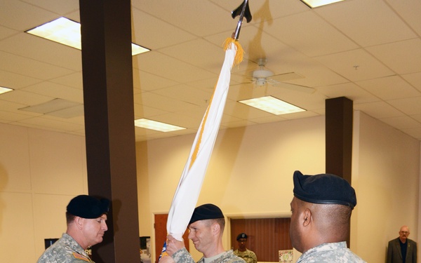 A U.S. Army change of command or activation ceremony taking place indoors, likely in a gymnasium or community center on a military installation. Two soldiers in the foreground are exchanging a furled guidon/colors on a staff, while a third soldier stands at attention observing. All personnel are wearing Army Combat Uniforms (ACU) with black berets.