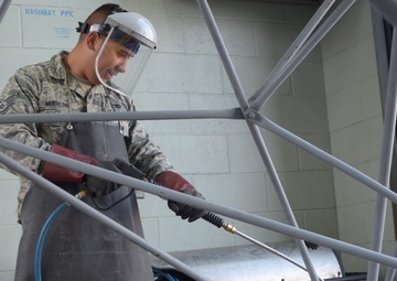 A U.S. Air Force service member identified by name tape as 'Muehling' is operating a Landa brand pressure washer in a designated washbay area. The individual is wearing full personal protective equipment including a face shield, protective apron, and gloves while cleaning what appears to be a geodesic or tubular metal frame structure. A sign reading 'WASHBAY PPE' is visible on the wall behind them.