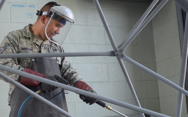 A U.S. Air Force service member identified by name tape as 'Muehling' is operating a Landa brand pressure washer in a designated washbay area. The individual is wearing full personal protective equipment including a face shield, protective apron, and gloves while cleaning what appears to be a geodesic or tubular metal frame structure. A sign reading 'WASHBAY PPE' is visible on the wall behind them.