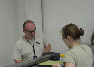 Two individuals in military-style attire are working together with what appears to be a sheet metal rolling or bending machine in an indoor workspace. Both are wearing safety glasses, suggesting a hands-on technical or fabrication task. A yellow sheet of material is being fed through the machine rollers.