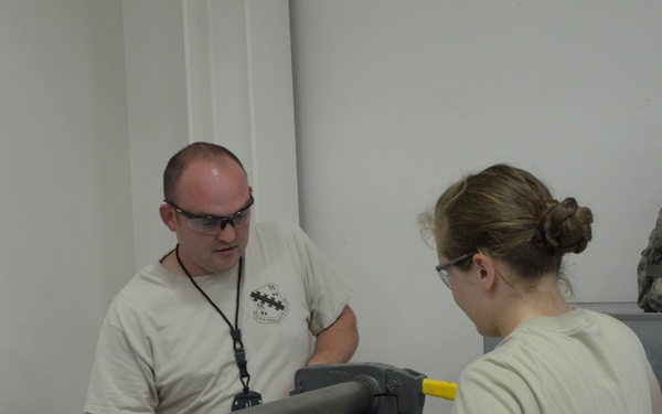 Two individuals in military-style attire are working together with what appears to be a sheet metal rolling or bending machine in an indoor workspace. Both are wearing safety glasses, suggesting a hands-on technical or fabrication task. A yellow sheet of material is being fed through the machine rollers.