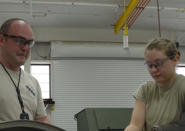 Two individuals wearing safety glasses and tan/olive military-style t-shirts are working together in what appears to be a military maintenance or fabrication shop. They are examining a document or technical manual alongside a curved metal component on a work surface. The setting suggests a training or maintenance environment with industrial equipment visible in the background.