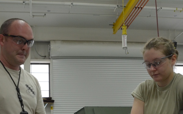 Two individuals wearing safety glasses and tan/olive military-style t-shirts are working together in what appears to be a military maintenance or fabrication shop. They are examining a document or technical manual alongside a curved metal component on a work surface. The setting suggests a training or maintenance environment with industrial equipment visible in the background.