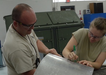 Two individuals wearing safety glasses and tan/khaki clothing are working together at a workbench in what appears to be a military or industrial workshop. The person on the right is marking a sheet metal panel with a green marker while the person on the left holds the panel steady. The environment contains various tools, equipment, and safety signage consistent with a metal fabrication or maintenance facility.
