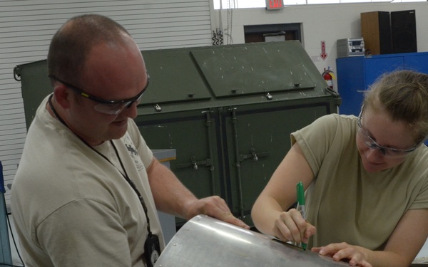 Two individuals wearing safety glasses and tan/khaki clothing are working together at a workbench in what appears to be a military or industrial workshop. The person on the right is marking a sheet metal panel with a green marker while the person on the left holds the panel steady. The environment contains various tools, equipment, and safety signage consistent with a metal fabrication or maintenance facility.