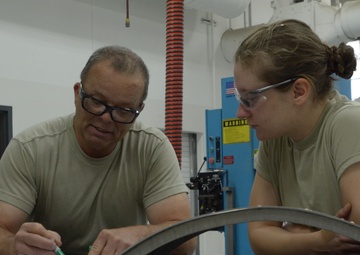 Two individuals wearing olive/tan military-style t-shirts are working together at a workbench in an industrial or military maintenance shop. The person on the left is writing on a document while both examine a curved metal component placed on the surface. The setting appears to be a fabrication or metalworking facility.